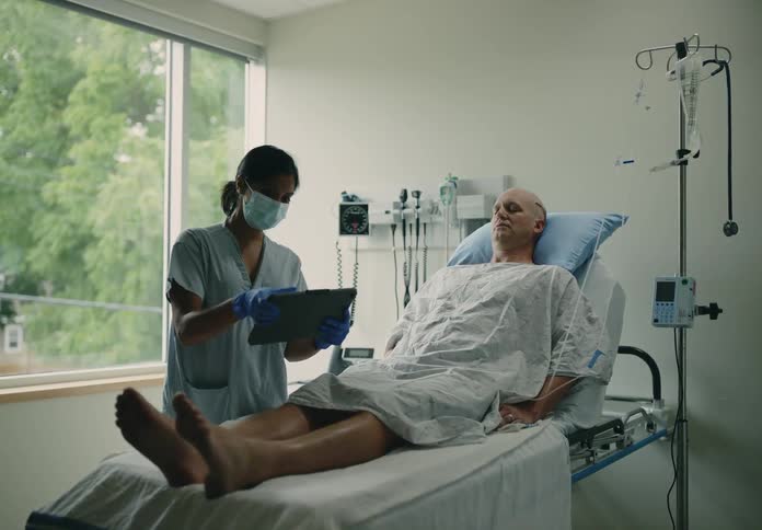 Patient with scar on head lying in hospital bed while the nurse wearing a face mask opens the window and pick up a tablet to show him