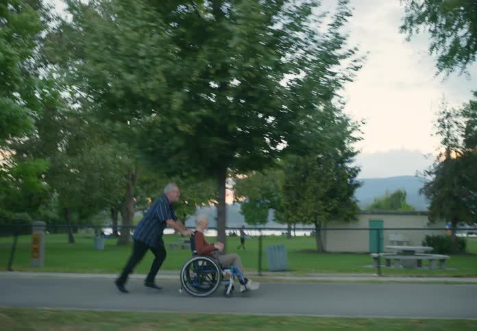 Middle aged man wearing shirt and jeans pushes woman in wheelchair by running next to public park