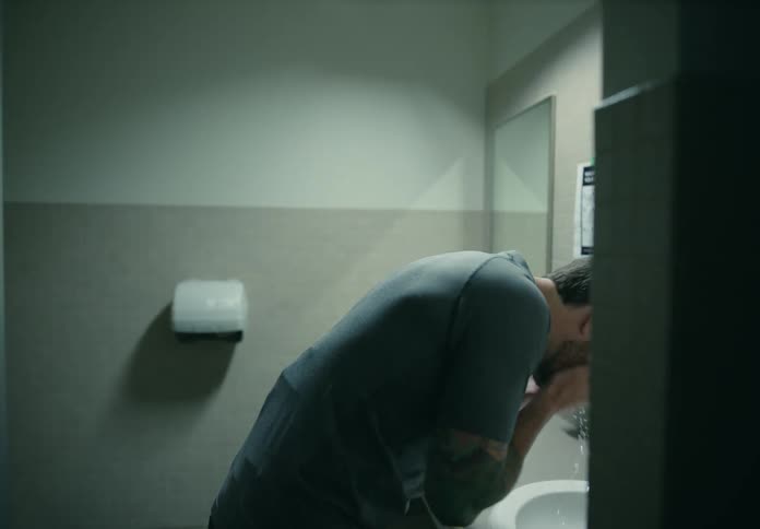 Distressed man facing the sink in a public restroom washing his face with water