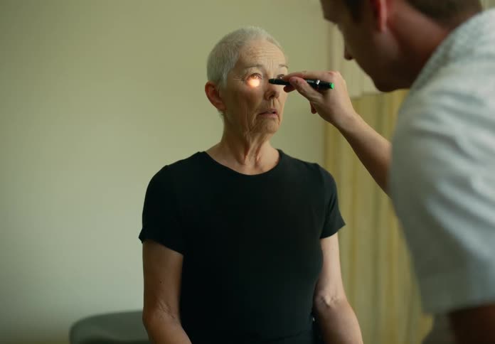 A woman is sitting in the doctors office and he shines a small flashlight in her eyes