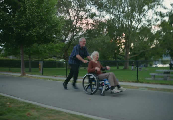 Running older man pushes woman in wheelchair through park with lake and red sky in the background