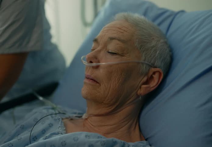 An elderly woman sleeps in a hospital bed as a nurse adjusts tubes