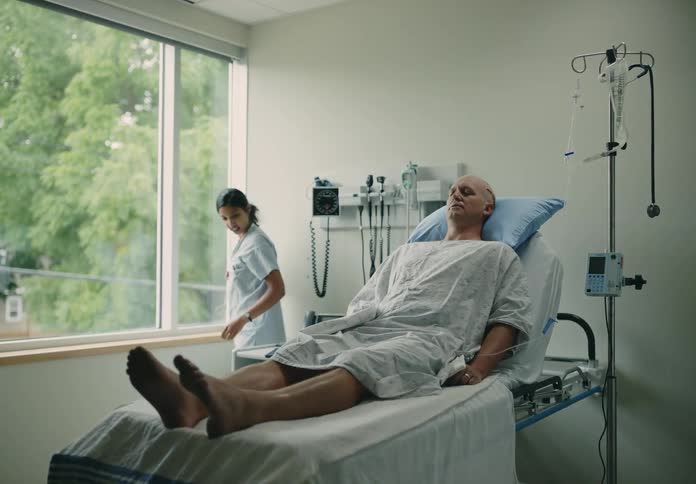 Patient with scar on head lying in hospital bed while the nurse opens the window and pick up a tablet to show him