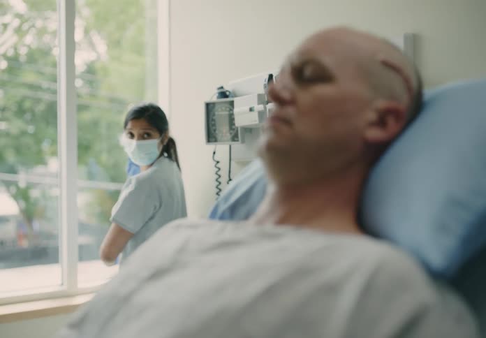 A nurse pulls up the window shade in the room as a patient lies in bed with a fresh surgical scar on his head
