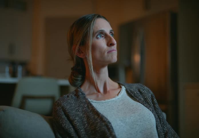 A woman is sitting down watching television when she hears a noise and gets up and walks across the room