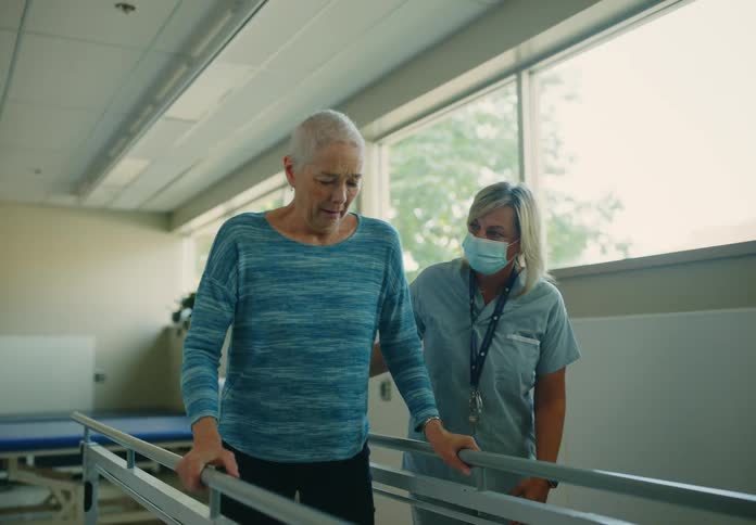 A woman attempts to walk using a set of walking rails in hospital as a nurse encourages her from the side
