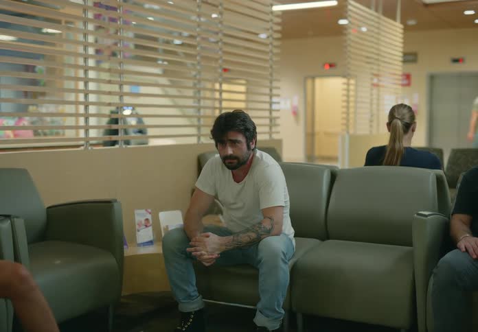 A man nervously sits on a couch in a hospital waiting room where other people also sit and wait
