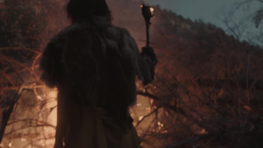 A man wearing clothes made of animal hide stands before a burning bonfire