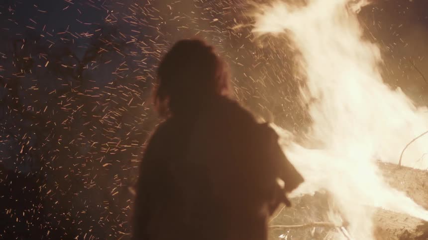 A man stands in front of a raging fire at night