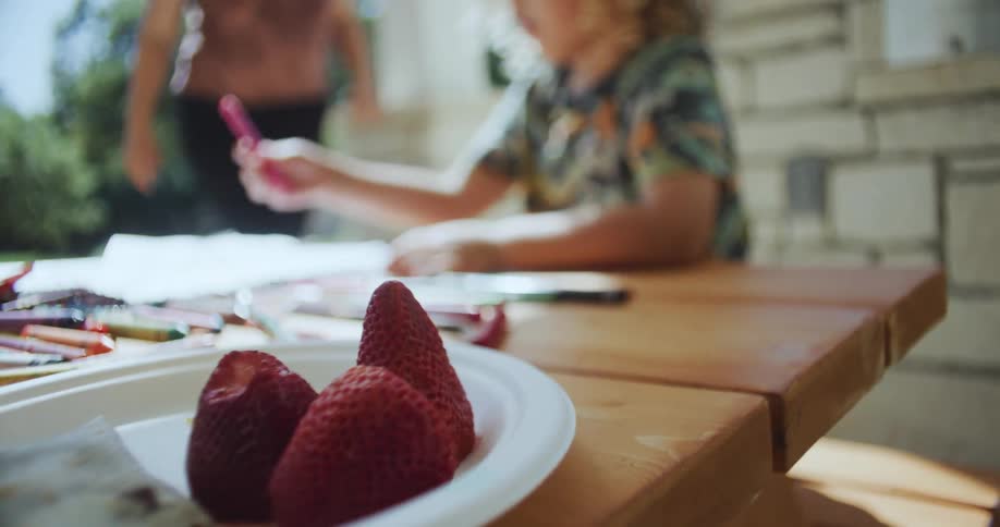 Blonde girl with curly hair and colorful shirt sits at table with strawberries on a plate drawing a picture with pink pencil in the backyard