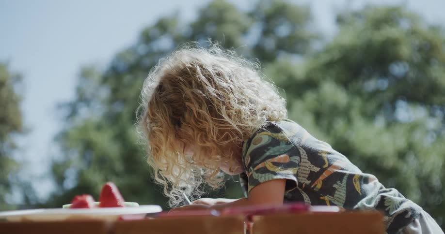 Blonde child with long curly hair climbs over a table on the backyard and starts drawing