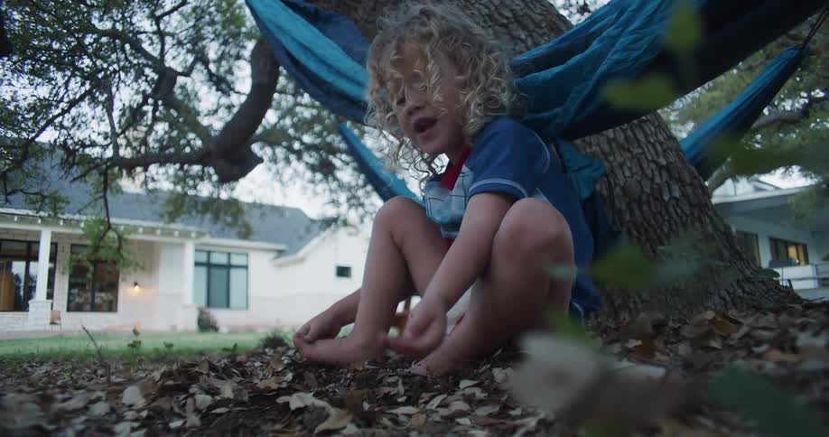Child sits on ground in yard of house playing with dirt and fallen leaves