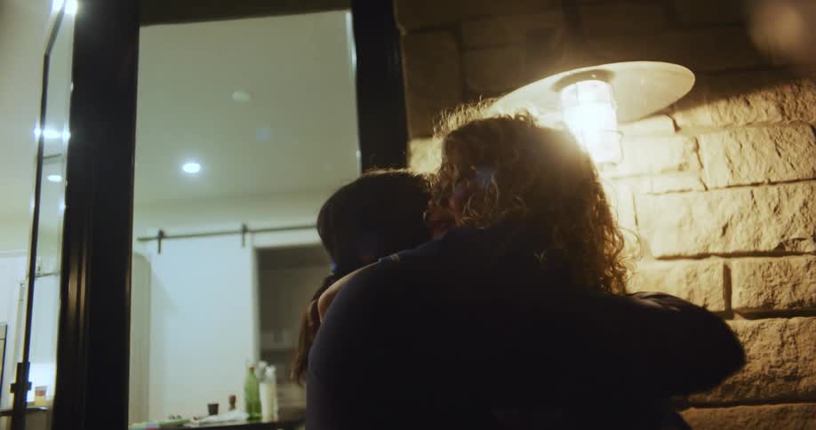 Mother holds daughter and they hug while talking outdoors near door to house at night