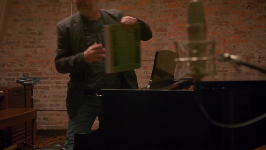 A man sitting at a piano stands up and takes his music book as he walks away