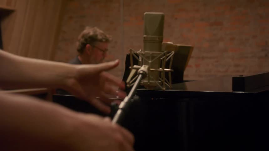 A man adjusts a microphone stand as an older man plays the piano nearby