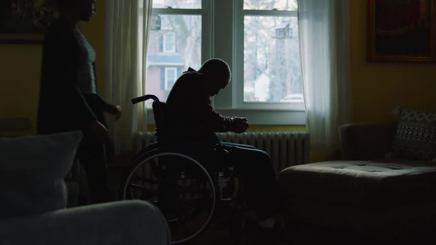 An elderly man sitting in a wheelchair in a dimly lit living room is approached and comforted by a woman