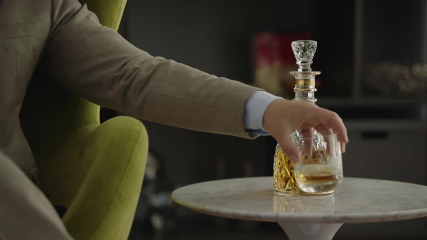 A man is sitting in a chair and he reaches for the glass of whisky on the table near a half full liquor decanter
