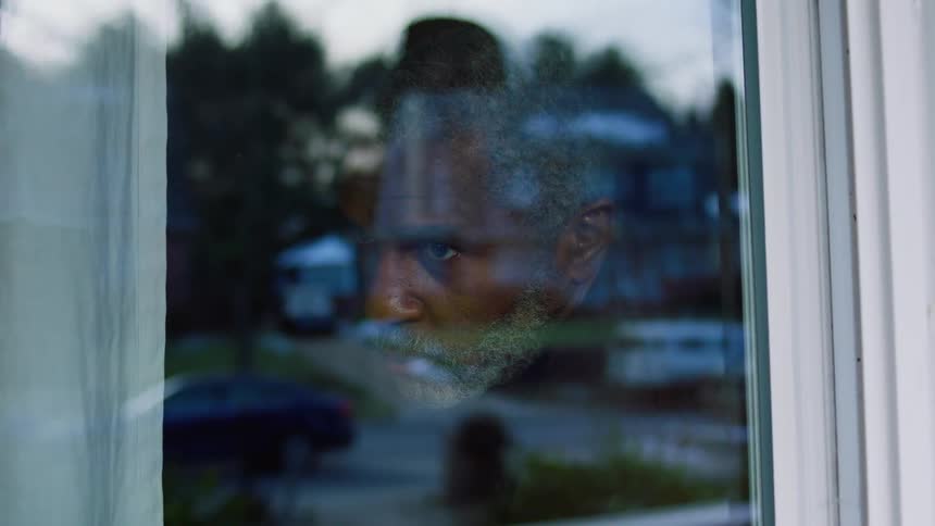 A man is inside his mouse looking out of the window and the reflection of passing cars is seen on that window