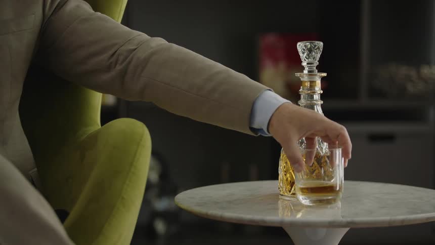 Man sitting in chair places drink glass on side table next to liquor carafe