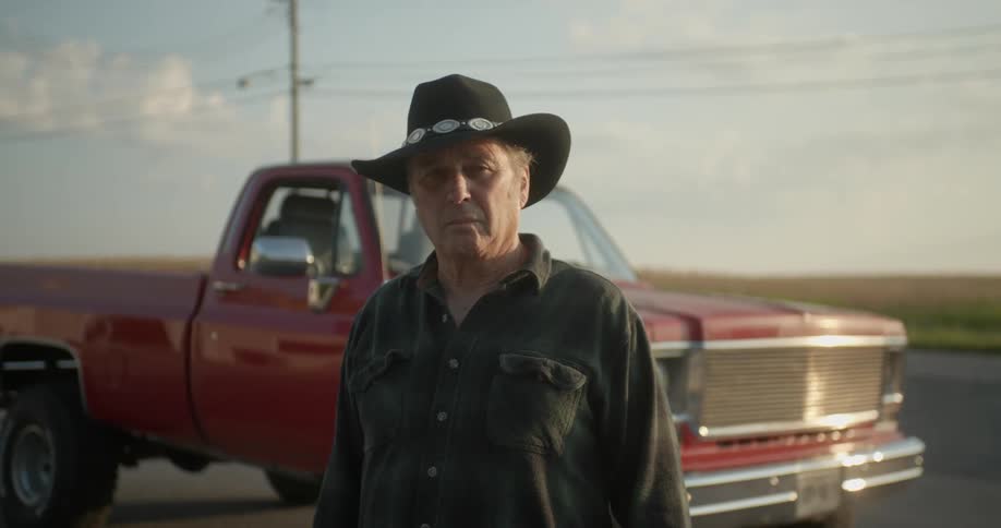 A man stands in front of his truck that is parked on the side of a road in front of a field and looks around