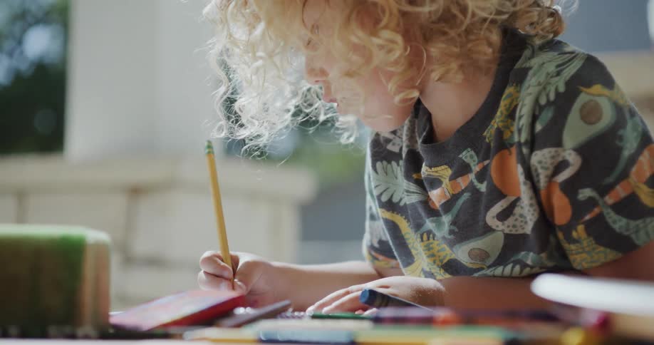 Blonde girl with curly hair and colorful shirt draws a picture with pencil sitting at table in the backyard