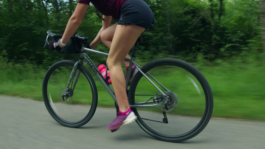 Female cyclist pedals bicycle along paved road near woods with mountains visible through the trees