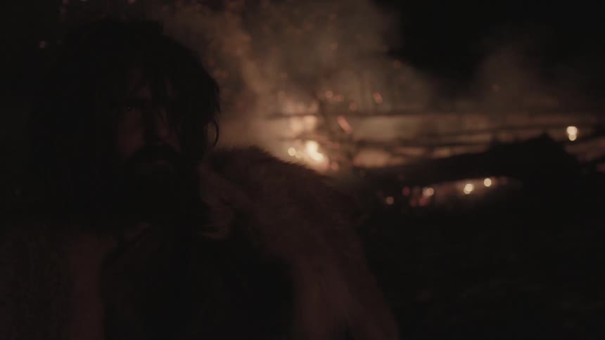 A man with shaggy hair wearing animal hides stands with a burning fire in the background