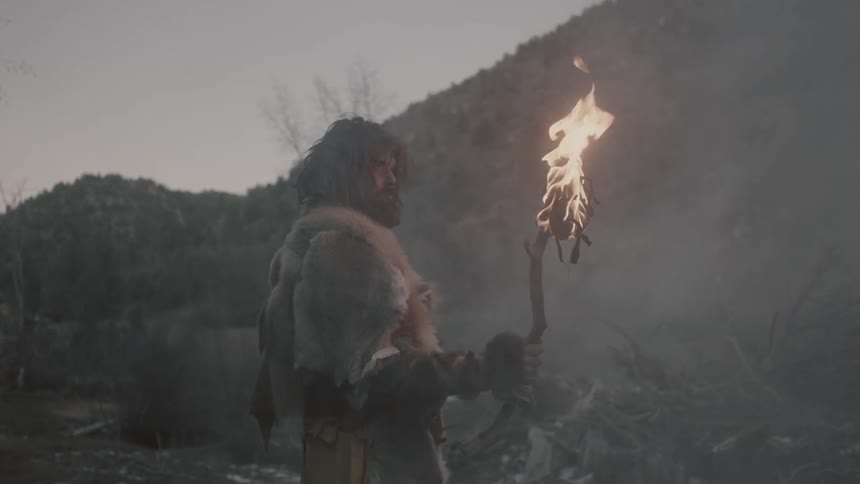 A man with shaggy hair and beard stands holding a burning torch