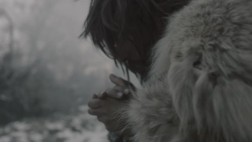Fur wearing man crouches while rubbing hands together in the cold