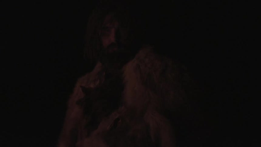 A man dressed in animal hides stands and looks upward in the darkness