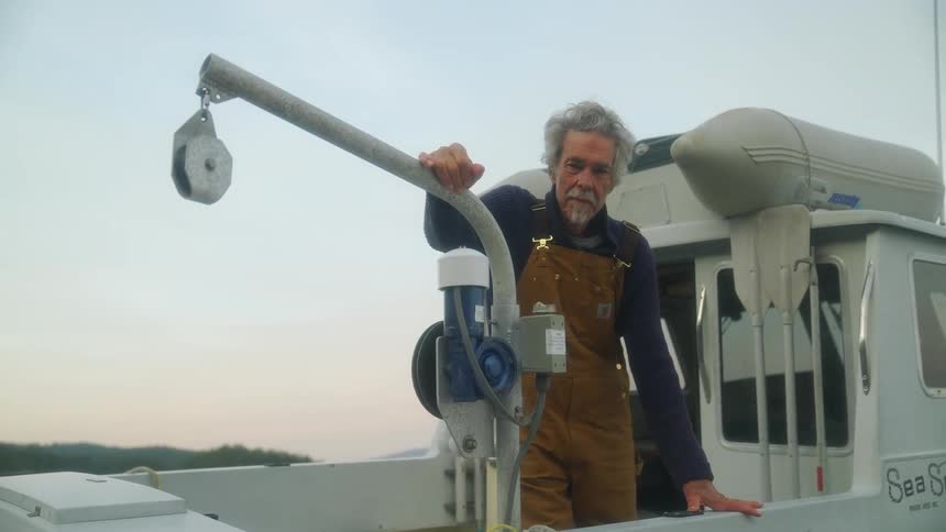 Man on bobbing boat leans against pot puller arm with forested shore in the background