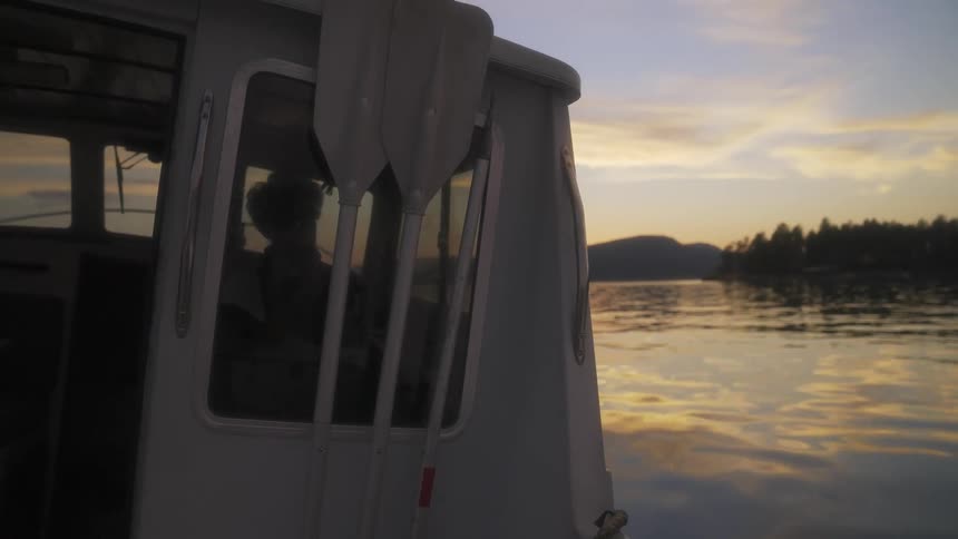 Person in pilot house steers boat over reflective water with forest and mountains on nearby shores