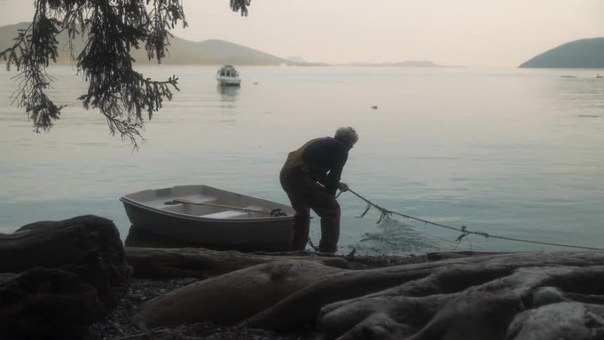 A man is standing along the shore pulling a rope tied to a small boat
