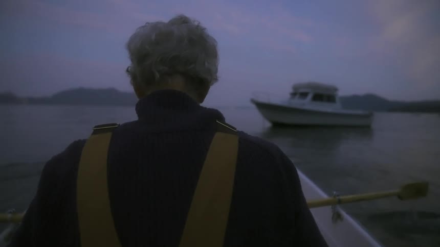 Back of an elderly fisherman rowing a rowboat near a boat on the calm surface of a lake at evening