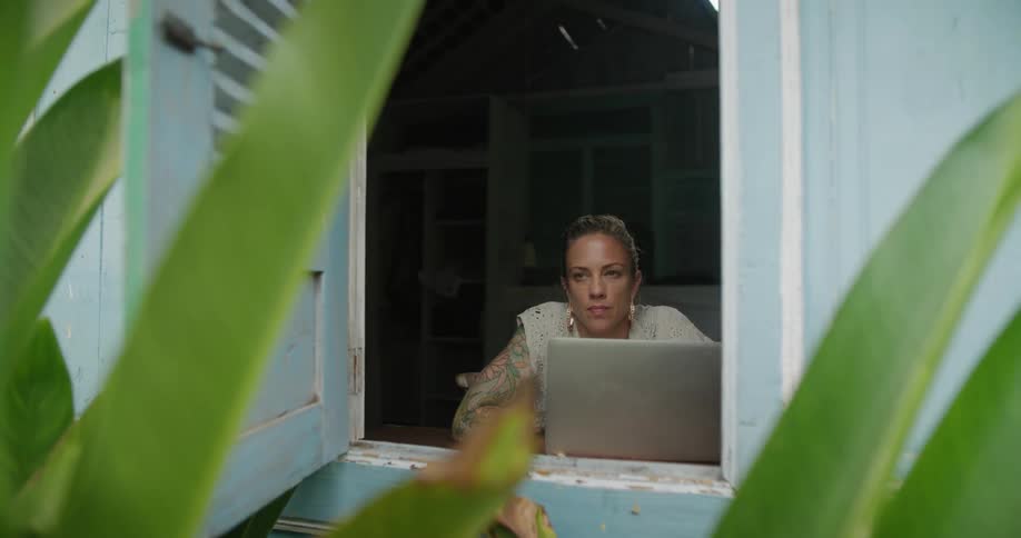 A woman types on her laptop while sitting at an open window