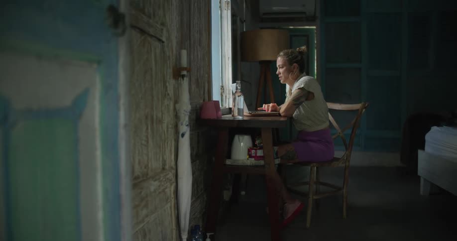 Woman with hoop earrings white shirt pink skirt and tattoos sits at desk uses laptop in bedroom