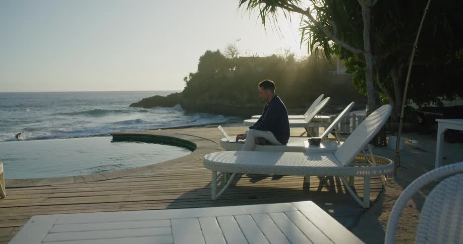 A man sits poolside at a resort and works on his laptop