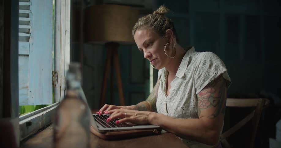 A woman types on a laptop while sitting at a desk in front of an open window during the day