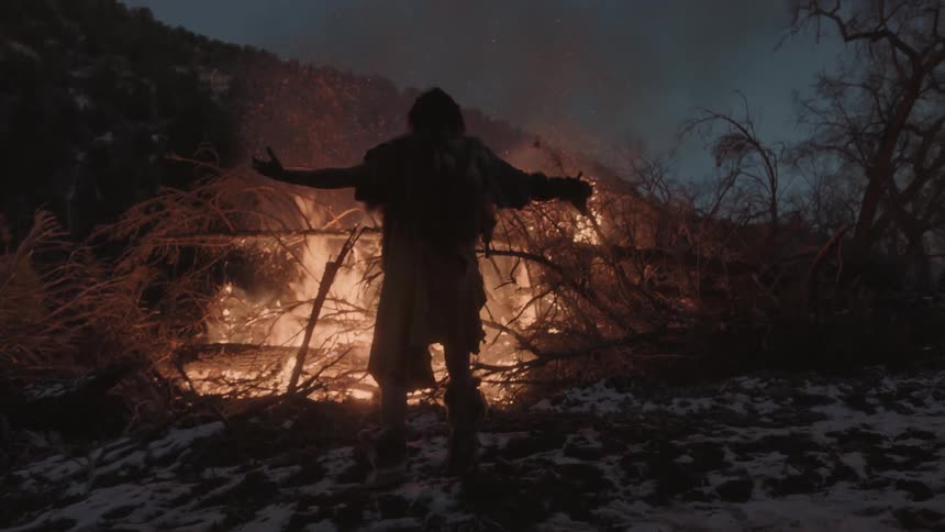 A man lifts his arms in front of a large fire at dawn