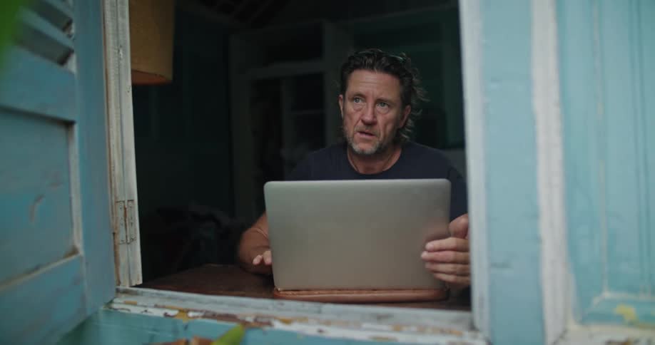 Mature man sitting near a window on a tropical house works on a laptop