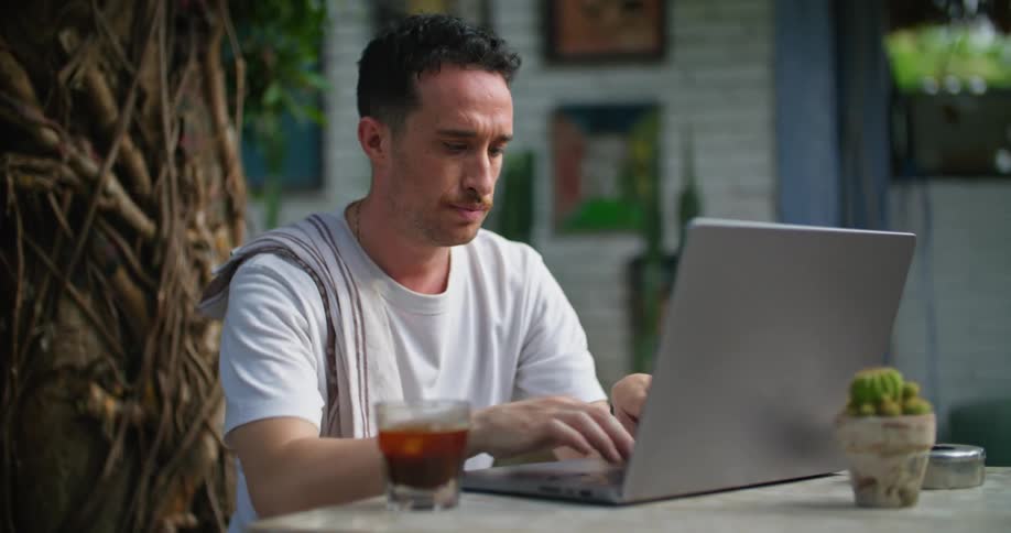 Sunlight is shining on the face of a man sitting outside at a table typing on a laptop computer