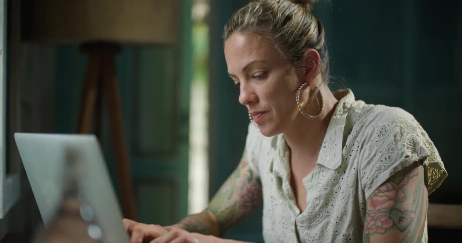 A woman with tattoos on her arms and bright pink nail polish is sitting at a table typing on a laptop computer