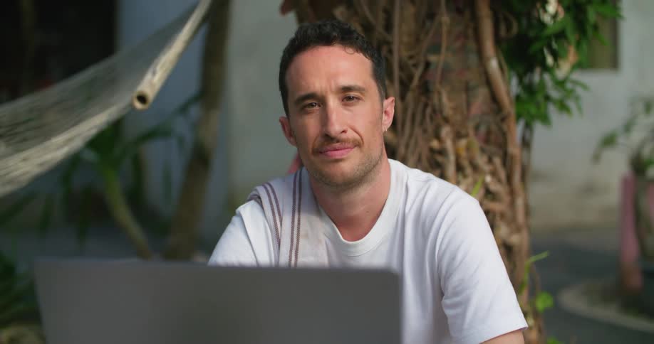 A man smiles while posing behind his laptop next to a hammock outside