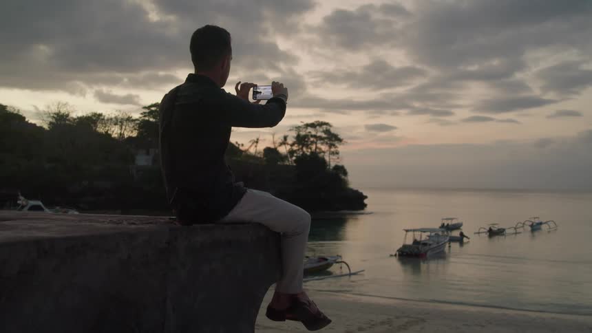 A man takes photos with his phone while sitting on the edge of a dock