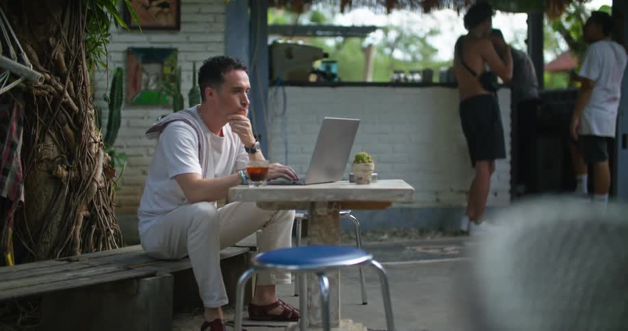 A man sits down at an outdoor restaurant with his laptop and a drink
