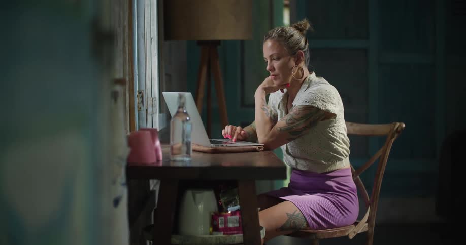 A woman is sitting at a table in a room typing on a laptop computer in front of a window