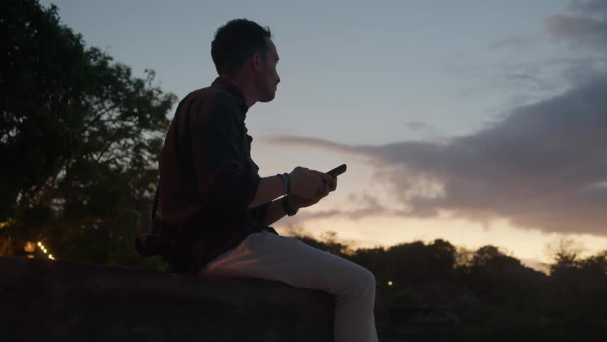 Man relaxes sitting on the countryside looking at his smartphone at evening