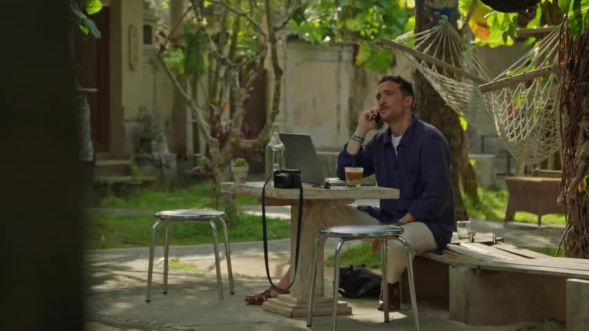 A man is sitting outside at a table with a laptop computer and talking on his phone