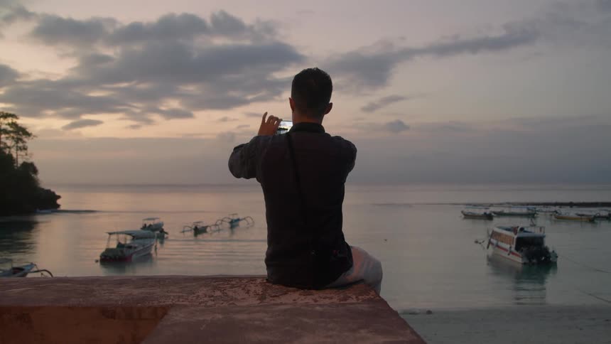 A man sitting on a wall overlooking the beach and marina uses his phone to photograph and video the scenery
