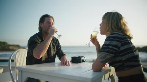 A couple toast and drink champagne while sitting outside at a seaside restaurant
